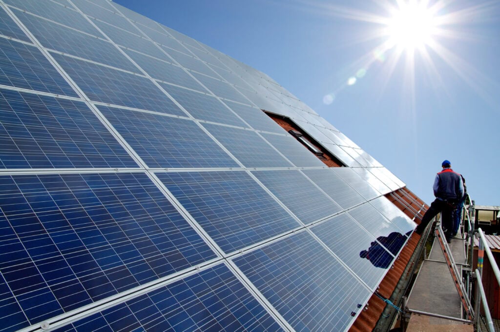 Techniker prüft Photovoltaik-Solarmodule auf einem Dach unter klarem Himmel, Sonnenlicht reflektiert auf den Modulen.