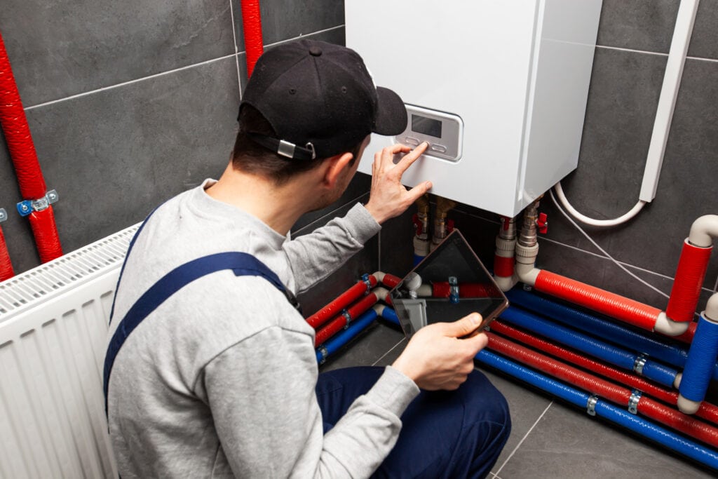 Techniker prüft eine Heizungsanlage in einem Installationsraum, neben roter und blauer Rohrleitung.